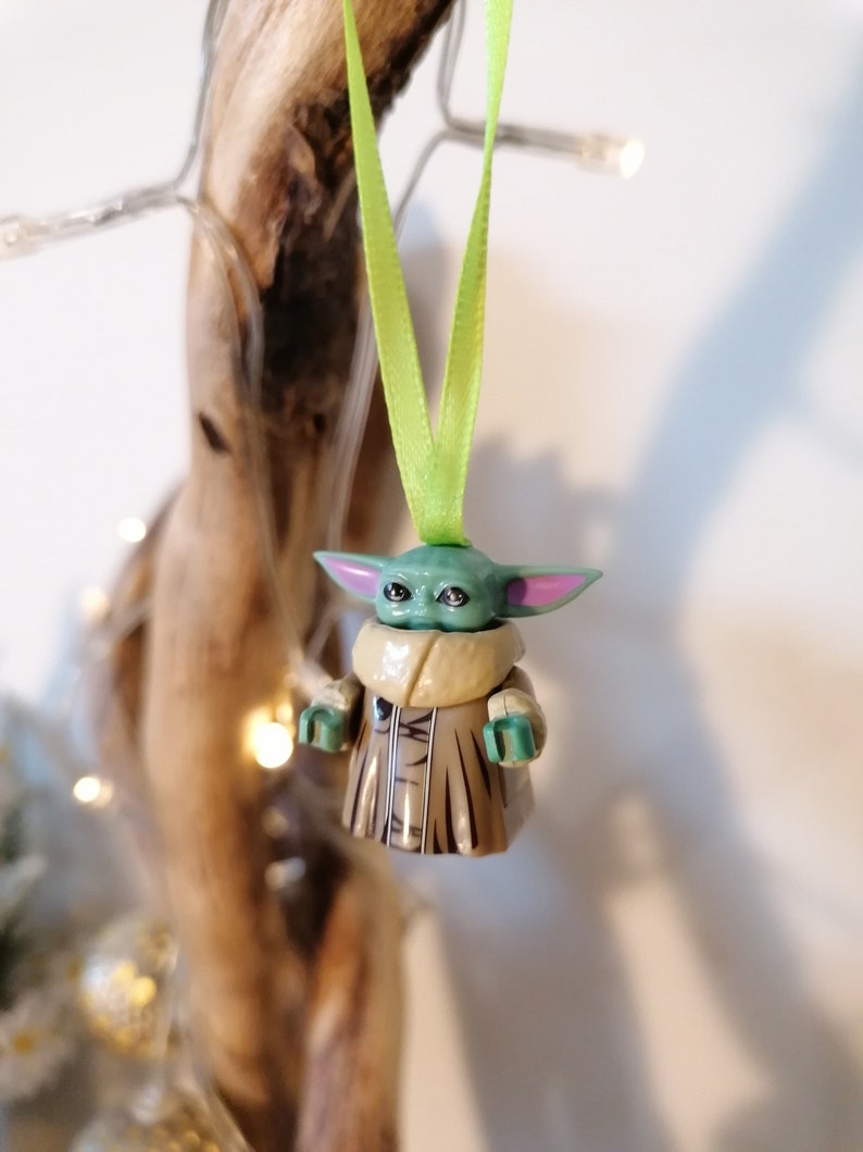 Puede incluir: Una figura de Lego verde y marr&oacute;n de un personaje de la franquicia Star Wars, colgada de una cinta verde. La figura lleva una t&uacute;nica marr&oacute;n y tiene orejas grandes.