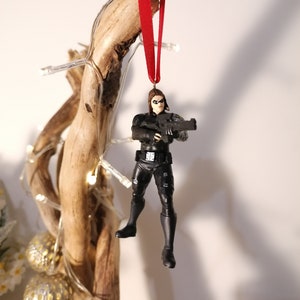 May include: A black and red Christmas ornament featuring a female action figure holding a gun. The figure is wearing a black outfit and has a red ribbon attached to the top of her head.