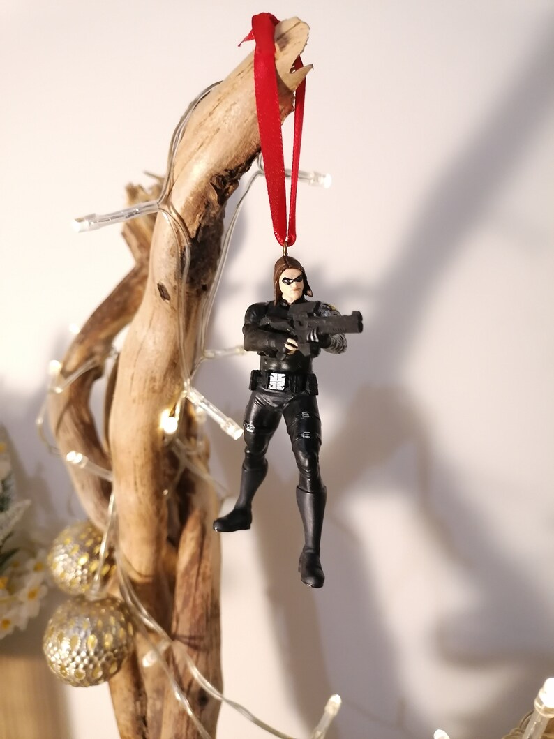 May include: A black and red Christmas ornament featuring a female action figure holding a gun. The figure is wearing a black outfit and has a red ribbon attached to the top of her head.