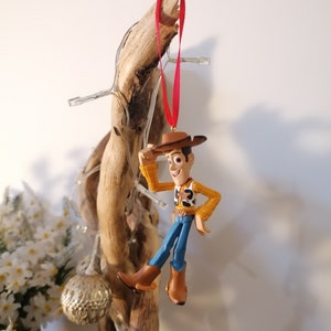 May include: A red and white Christmas bauble featuring a plastic figurine of Woody from the Toy Story film franchise. Woody is wearing his signature cowboy hat, waistcoat, and jeans. He is holding his hat in his right hand and smiling.