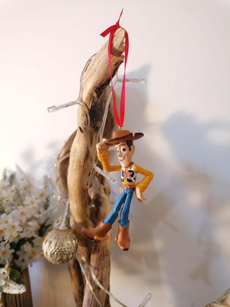 May include: A red and white Christmas bauble featuring a plastic figurine of Woody from the Toy Story film franchise. Woody is wearing his signature cowboy hat, waistcoat, and jeans. He is holding his hat in his right hand and smiling.