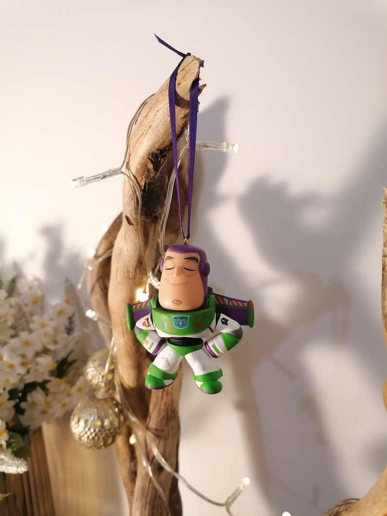 K&ouml;nnte beinhalten: Ein gr&uuml;ner und lilafarbener Plastik-Anh&auml;nger von Buzz Lightyear aus der Toy Story-Reihe. Der Anh&auml;nger h&auml;ngt an einem lilafarbenen Band an einem St&uuml;ck Treibholz.