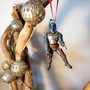 May include: A blue and gray Star Wars Boba Fett action figure ornament hanging from a driftwood branch with silver and white decorative balls. The ornament is attached to a red ribbon.
