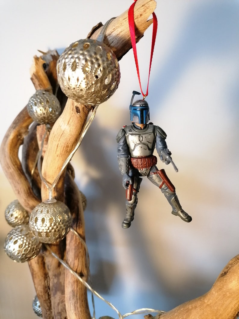 May include: A blue and gray Star Wars Boba Fett action figure ornament hanging from a driftwood branch with silver and white decorative balls. The ornament is attached to a red ribbon.
