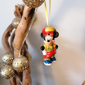 May include: A red, yellow and green Mickey Mouse ornament with a red hat and blue shoes. The ornament is hanging from a yellow ribbon.