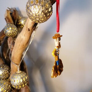 May include: A red string ornament with a small plastic figurine of Woody from Toy Story. Woody is wearing his signature cowboy hat, vest, and boots. He is standing on a small brown base.
