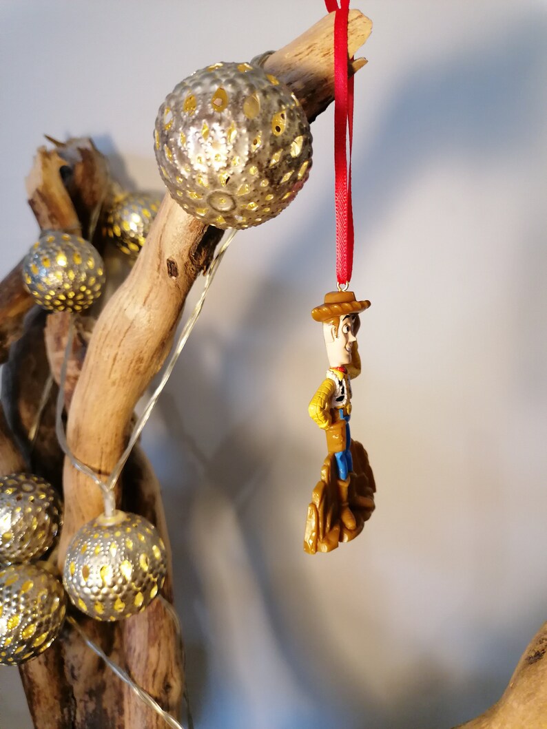 May include: A red string ornament with a small plastic figurine of Woody from Toy Story. Woody is wearing his signature cowboy hat, vest, and boots. He is standing on a small brown base.