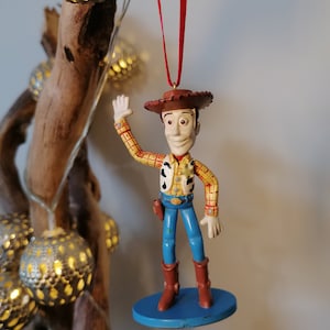 May include: A plastic figurine of Woody from Toy Story, wearing his signature cowboy hat, vest, and jeans. He is waving with his right hand and has a friendly expression on his face. The figurine is hanging from a red ribbon.