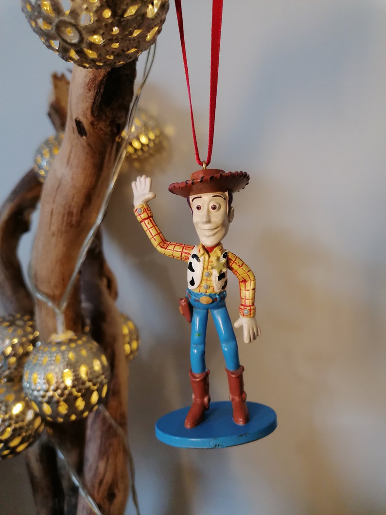 May include: A plastic figurine of Woody from Toy Story, wearing his signature cowboy hat, vest, and jeans. He is waving with his right hand and has a friendly expression on his face. The figurine is hanging from a red ribbon.