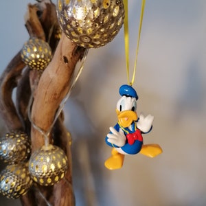 Op de afbeelding: Een gele draad hangt aan een tak met een blauw en geel Donald Duck ornament. Het ornament is een 3D-figuur van Donald Duck met een blauwe matrozenpet en een rode strik. De tak is versierd met zilveren en gouden kerstversieringen.