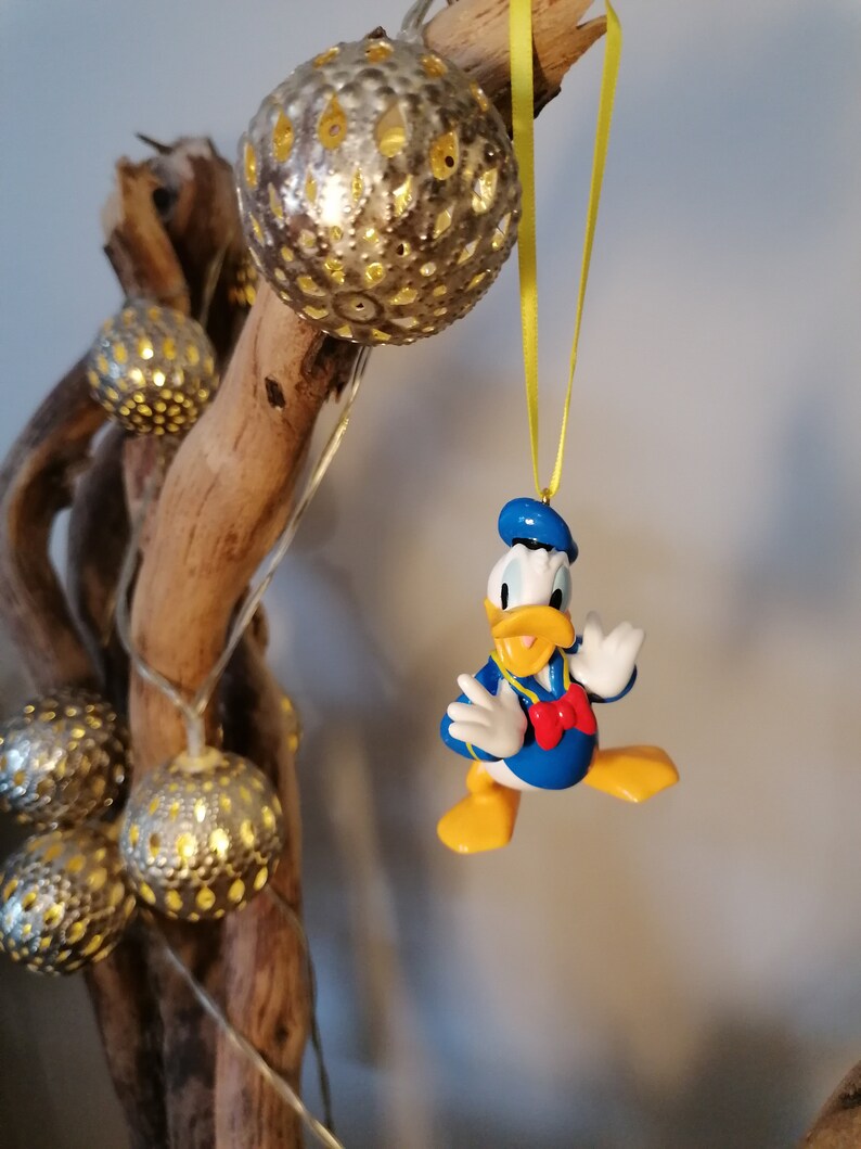 Op de afbeelding: Een gele draad hangt aan een tak met een blauw en geel Donald Duck ornament. Het ornament is een 3D-figuur van Donald Duck met een blauwe matrozenpet en een rode strik. De tak is versierd met zilveren en gouden kerstversieringen.