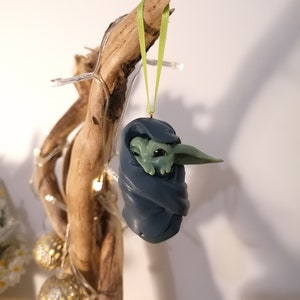 May include: A green and blue Christmas ornament featuring a character from the Star Wars franchise, wrapped in a blue blanket. The ornament is hanging from a green ribbon on a branch.