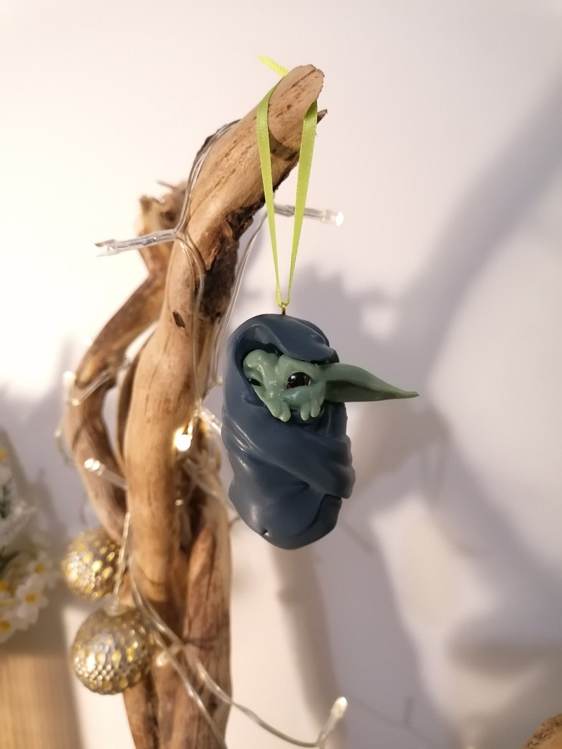 May include: A green and blue Christmas ornament featuring a character from the Star Wars franchise, wrapped in a blue blanket. The ornament is hanging from a green ribbon on a branch.