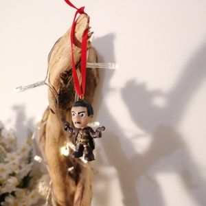 May include: A small, dark-haired figurine of a man wearing a brown and black outfit, holding two guns, is hanging from a piece of driftwood with a red ribbon.