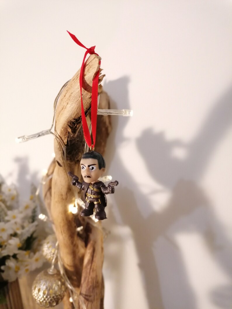 May include: A small, dark-haired figurine of a man wearing a brown and black outfit, holding two guns, is hanging from a piece of driftwood with a red ribbon.