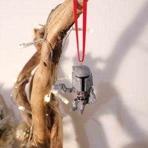 May include: A grey and brown Mandalorian Christmas ornament with a red ribbon hanging from a branch.