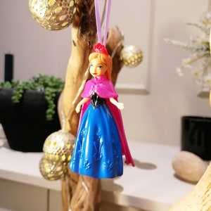 May include: A plastic doll ornament of a princess wearing a blue dress and a pink cape. The doll is hanging from a purple string.
