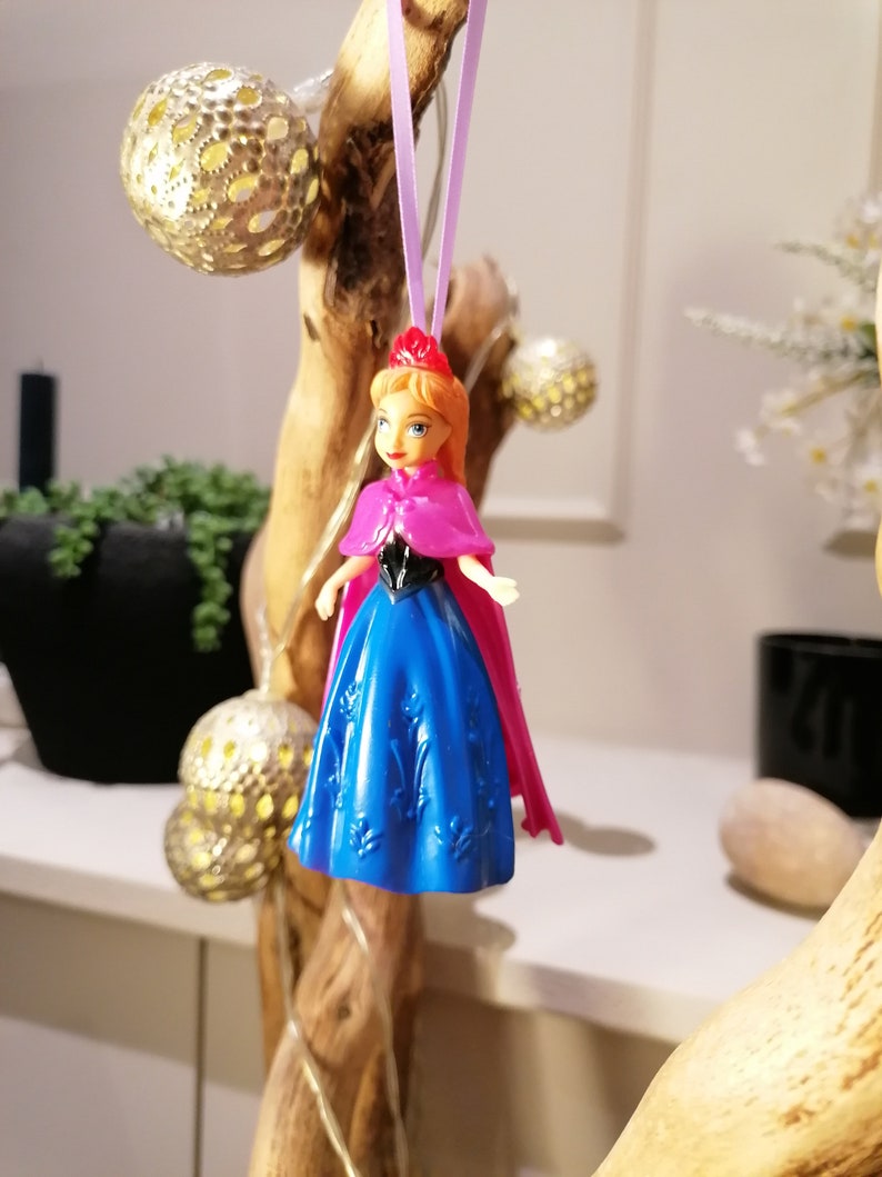 May include: A plastic doll ornament of a princess wearing a blue dress and a pink cape. The doll is hanging from a purple string.