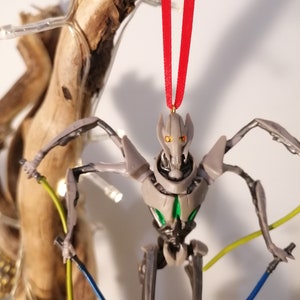 May include: A gray and green robot action figure with red eyes and a red ribbon hanging from its head. The robot is holding two lightsabers, one yellow and one blue.