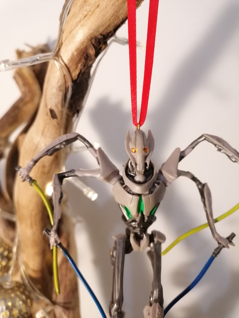 May include: A gray and green robot action figure with red eyes and a red ribbon hanging from its head. The robot is holding two lightsabers, one yellow and one blue.