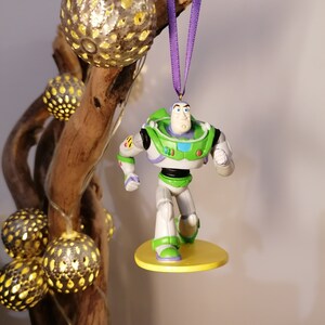 Puede incluir: Una figura de plástico verde y blanca de Buzz Lightyear de la franquicia de películas Toy Story colgada de una cinta morada. La figura está de pie sobre una base amarilla.