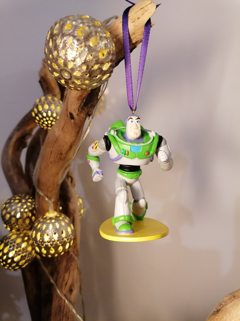 K&ouml;nnte beinhalten: Eine gr&uuml;ne und wei&szlig;e Plastikfigur von Buzz Lightyear aus der Toy Story-Filmreihe, die an einem violetten Band h&auml;ngt. Die Figur steht auf einem gelben Sockel.