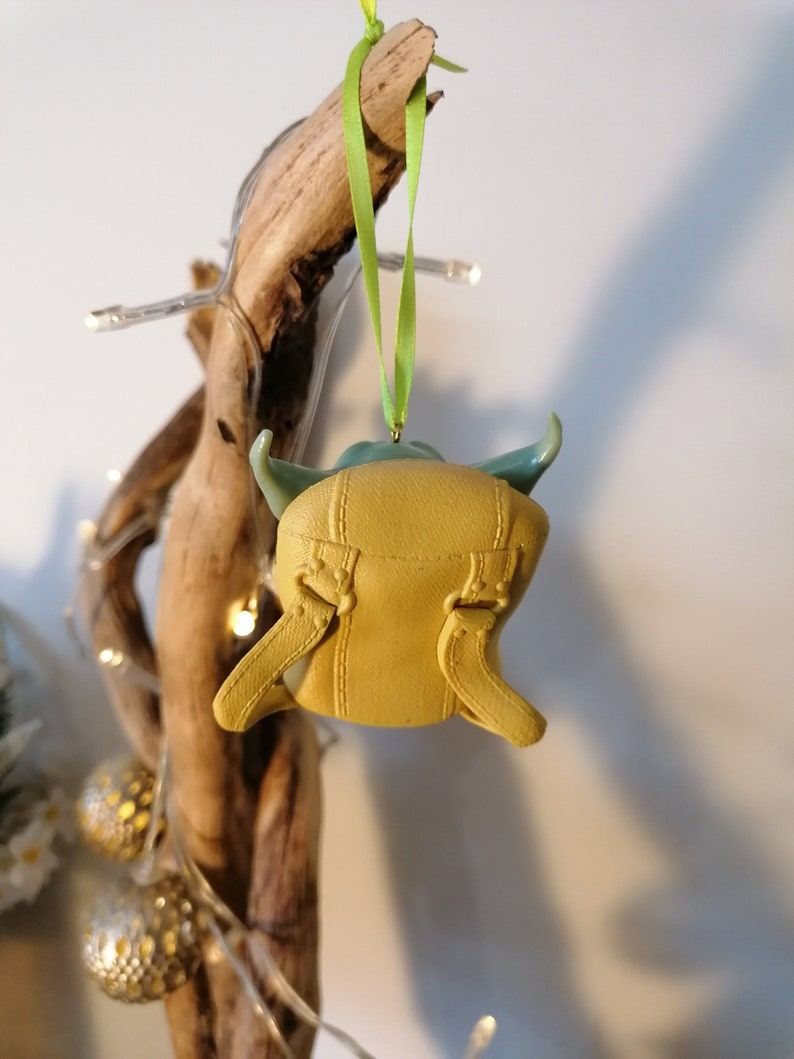 May include: A yellow and green Christmas ornament shaped like a character from the Star Wars franchise. The ornament is hanging from a green ribbon.