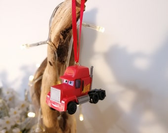 Op de afbeelding: Rode vrachtwagen kerstversiering met een glimlachend gezicht en het woord "Mater" aan de zijkant. De versiering hangt aan een rood lint.
