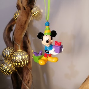 Peut inclure: Une d&eacute;coration de No&euml;l repr&eacute;sentant Mickey Mouse, color&eacute;, avec un chapeau de f&ecirc;te et tenant une bo&icirc;te cadeau violette. La d&eacute;coration est suspendue &agrave; un ruban vert.