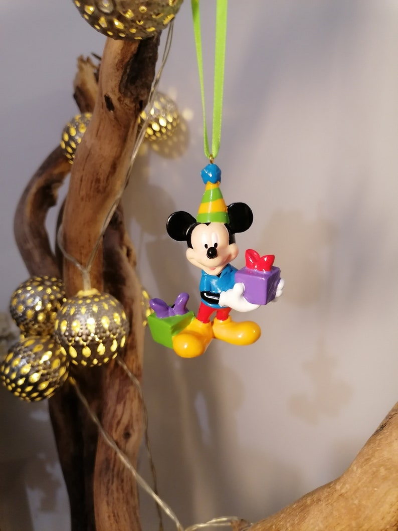 Peut inclure: Une d&eacute;coration de No&euml;l repr&eacute;sentant Mickey Mouse, color&eacute;, avec un chapeau de f&ecirc;te et tenant une bo&icirc;te cadeau violette. La d&eacute;coration est suspendue &agrave; un ruban vert.