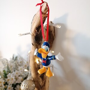 May include: A blue and yellow Donald Duck Christmas ornament with a red ribbon hanging from a piece of driftwood.