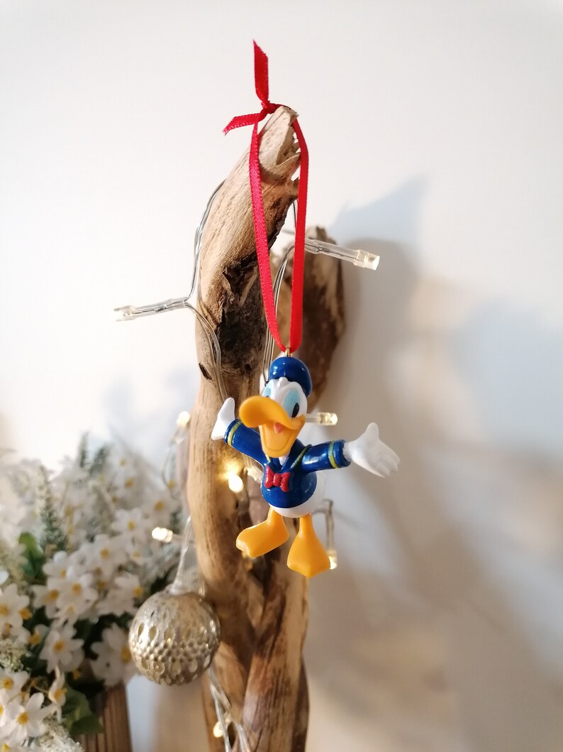 May include: A blue and yellow Donald Duck Christmas ornament with a red ribbon hanging from a piece of driftwood.