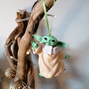 May include: A green and beige Christmas ornament featuring a character from the Star Wars franchise, The Mandalorian. The ornament is hanging from a branch with green ribbon.
