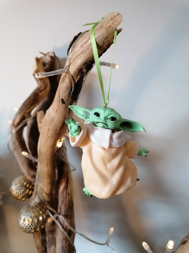May include: A green and beige Christmas ornament featuring a character from the Star Wars franchise, The Mandalorian. The ornament is hanging from a branch with green ribbon.