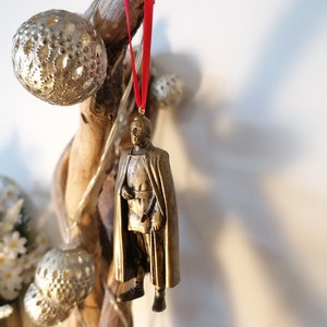 May include: A gold-colored ornament depicting a person wearing a cape and holding a staff. The ornament is hanging from a red ribbon.
