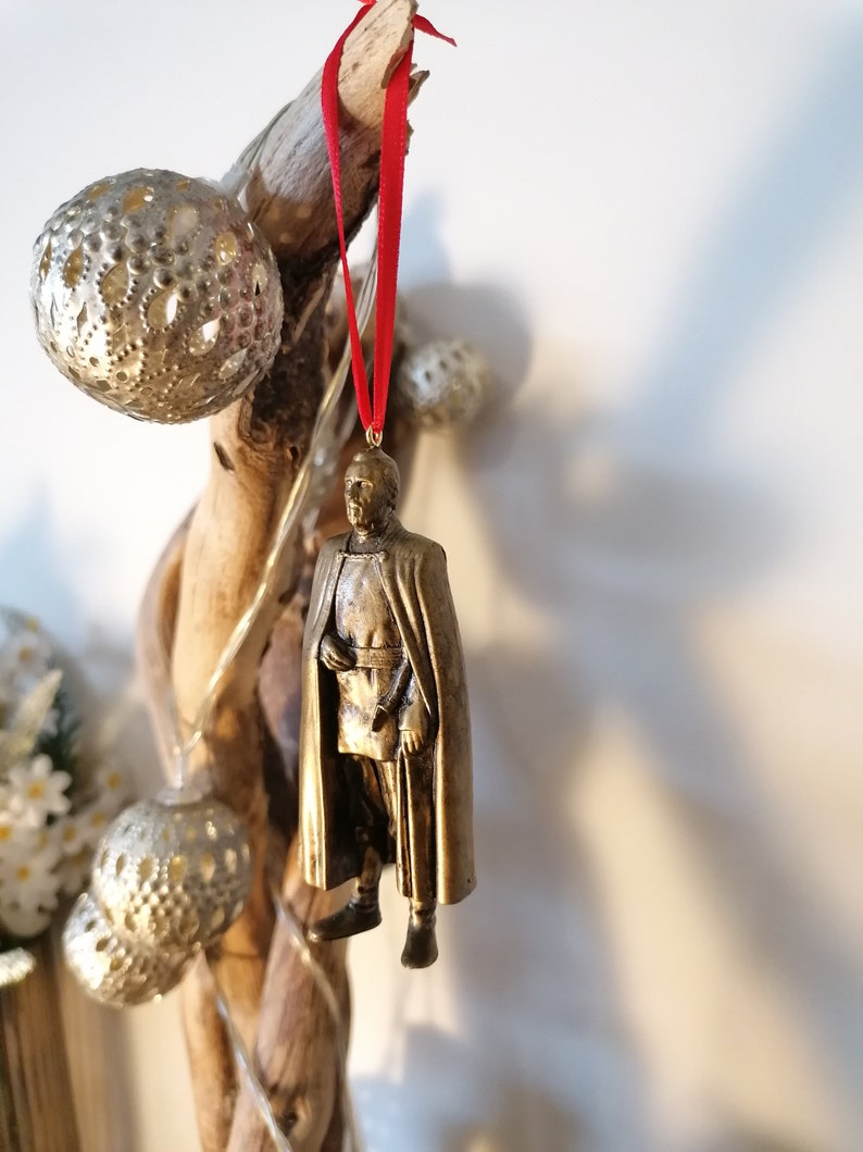 May include: A gold-colored ornament depicting a person wearing a cape and holding a staff. The ornament is hanging from a red ribbon.