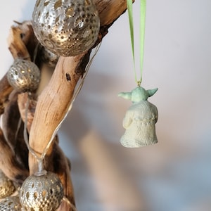 May include: A green Yoda Christmas ornament hanging from a branch with a green ribbon. The ornament is made of ceramic and has a detailed design.