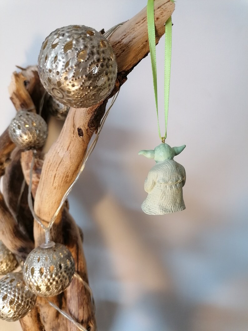 May include: A green Yoda Christmas ornament hanging from a branch with a green ribbon. The ornament is made of ceramic and has a detailed design.
