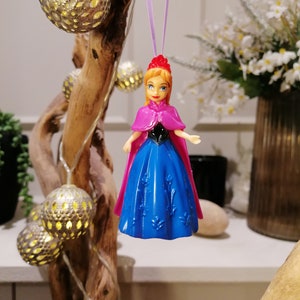 May include: A plastic doll wearing a blue dress and a pink cape, hanging from a purple string. The doll has a red crown and brown hair. The doll is hanging in front of a wooden branch with string lights.