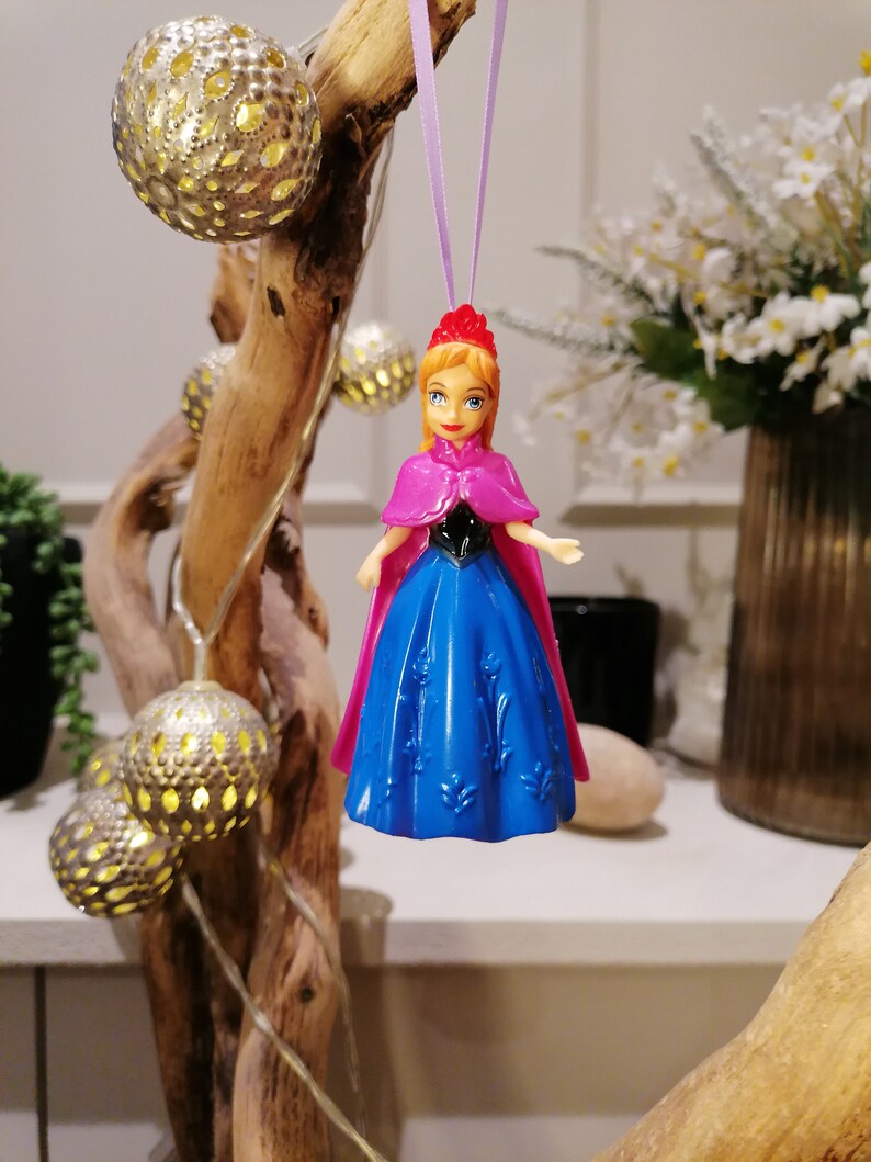 May include: A plastic doll wearing a blue dress and a pink cape, hanging from a purple string. The doll has a red crown and brown hair. The doll is hanging in front of a wooden branch with string lights.