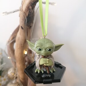 Puede incluir: Adorno de Navidad de Star Wars Yoda verde con una base hexagonal negra y una cinta verde.