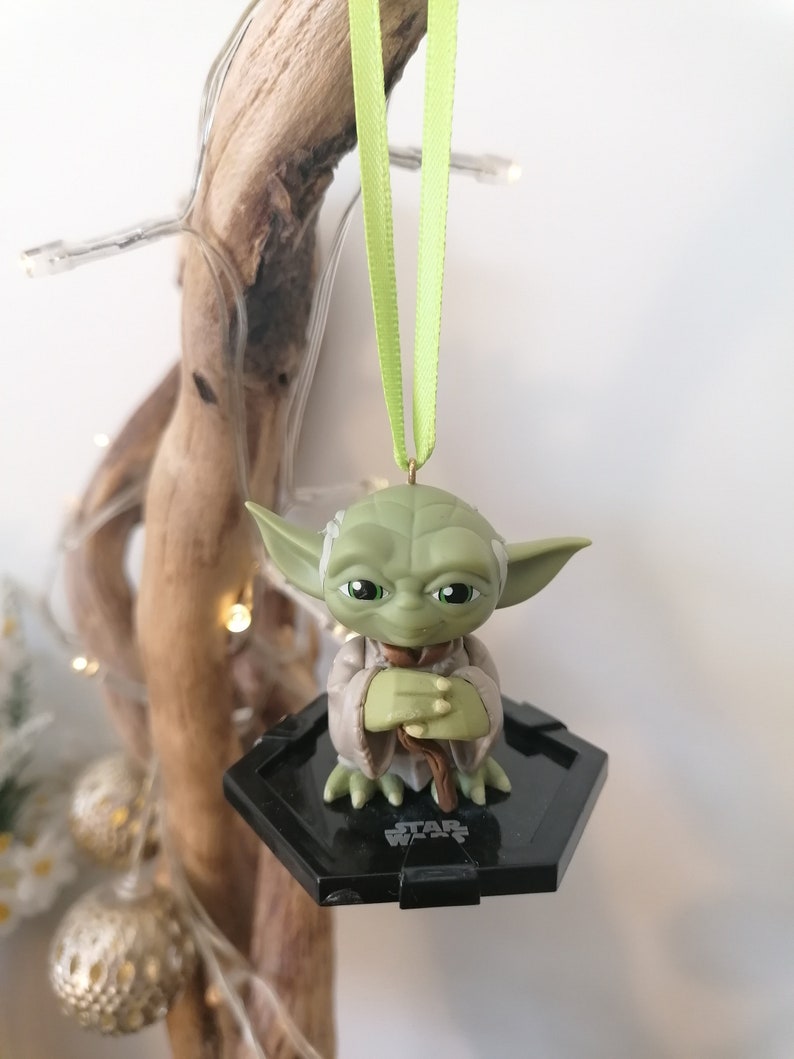 Puede incluir: Adorno de Navidad de Star Wars Yoda verde con una base hexagonal negra y una cinta verde.