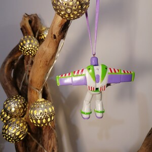 Peut inclure: Une d&eacute;coration de No&euml;l Buzz l'&Eacute;clair est suspendue &agrave; un ruban violet. L'ornement est blanc, vert et violet. Des guirlandes lumineuses argent&eacute;es d&eacute;coratives avec des lumi&egrave;res jaunes sont suspendues &agrave; un morceau de bois flott&eacute;.