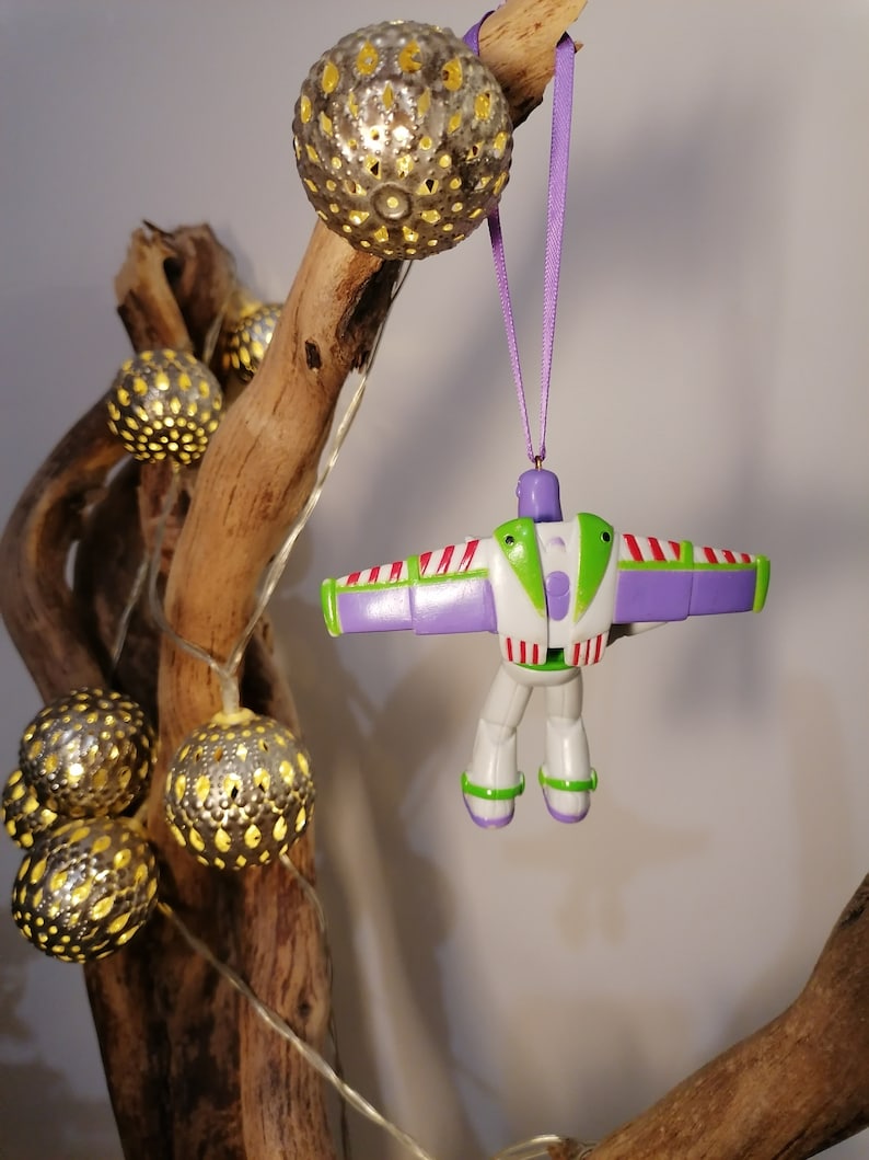 Peut inclure: Une d&eacute;coration de No&euml;l Buzz l'&Eacute;clair est suspendue &agrave; un ruban violet. L'ornement est blanc, vert et violet. Des guirlandes lumineuses argent&eacute;es d&eacute;coratives avec des lumi&egrave;res jaunes sont suspendues &agrave; un morceau de bois flott&eacute;.