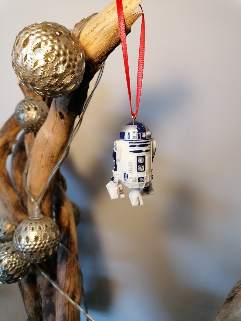 K&ouml;nnte beinhalten: Ein wei&szlig;er und blauer R2-D2 Star Wars Charakter Anh&auml;nger, der an einem roten Band an einem Treibholzast h&auml;ngt.
