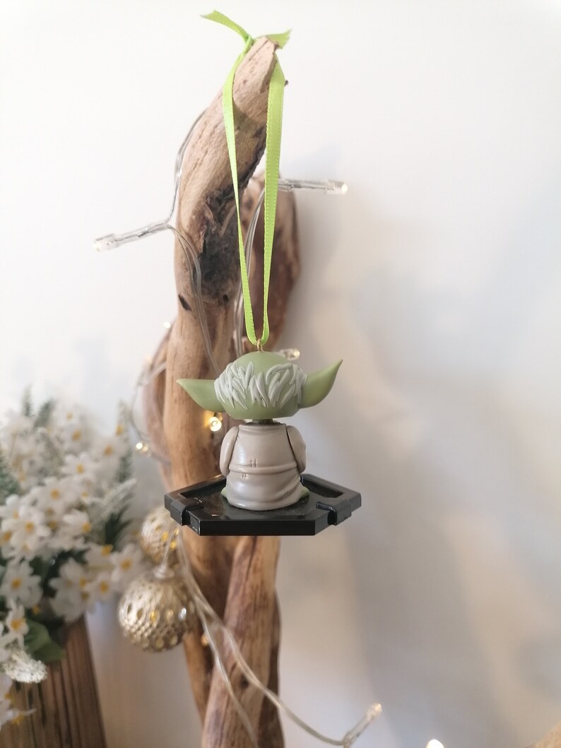 Puede incluir: Un adorno de Yoda verde y marr&oacute;n colgado de una rama con una cinta verde. El adorno es una peque&ntilde;a figura de Yoda con una base negra.