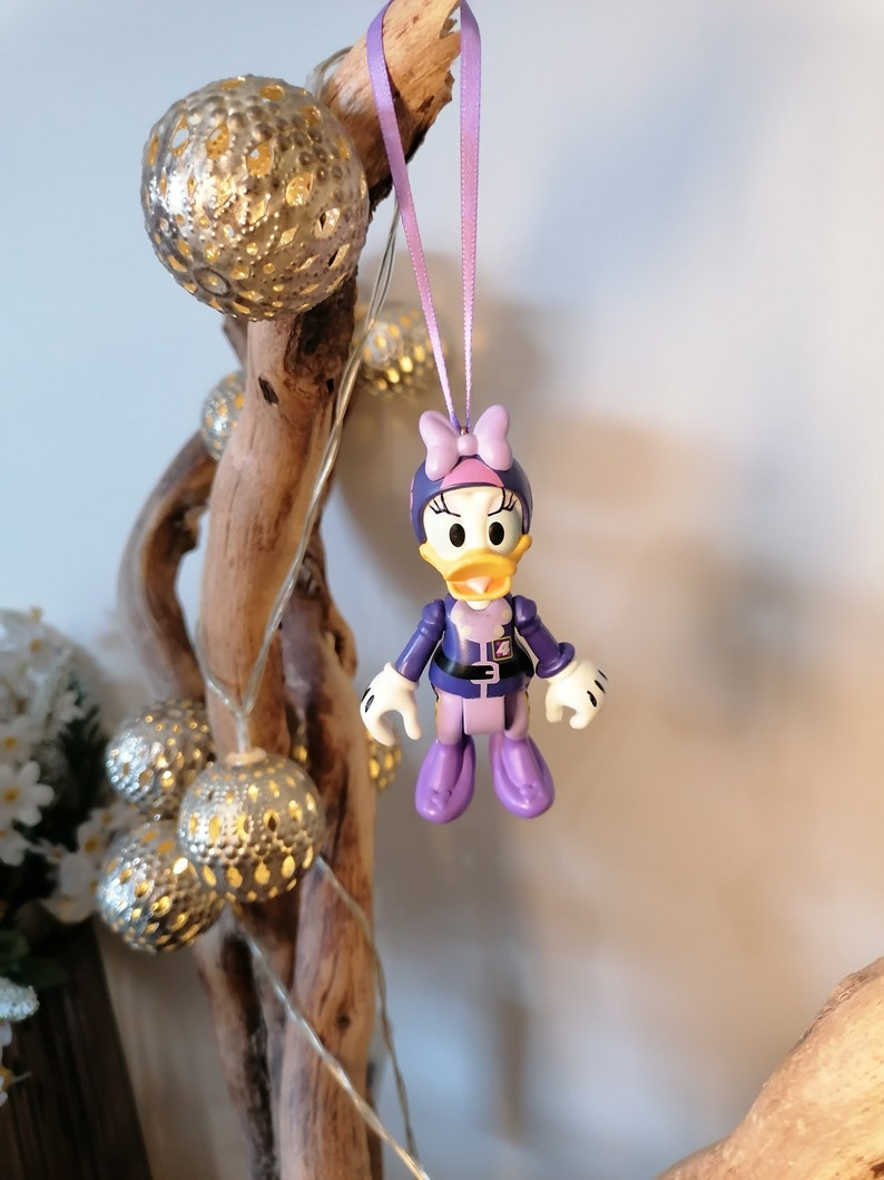 May include: Purple and yellow plastic figurine of a duck wearing a purple and white outfit with a pink bow. The figurine is hanging from a purple ribbon.