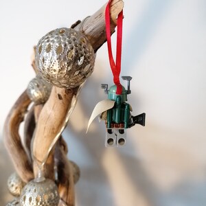 May include: A green and brown Lego Star Wars Boba Fett ornament with a red ribbon hanging from a branch with silver ornaments.