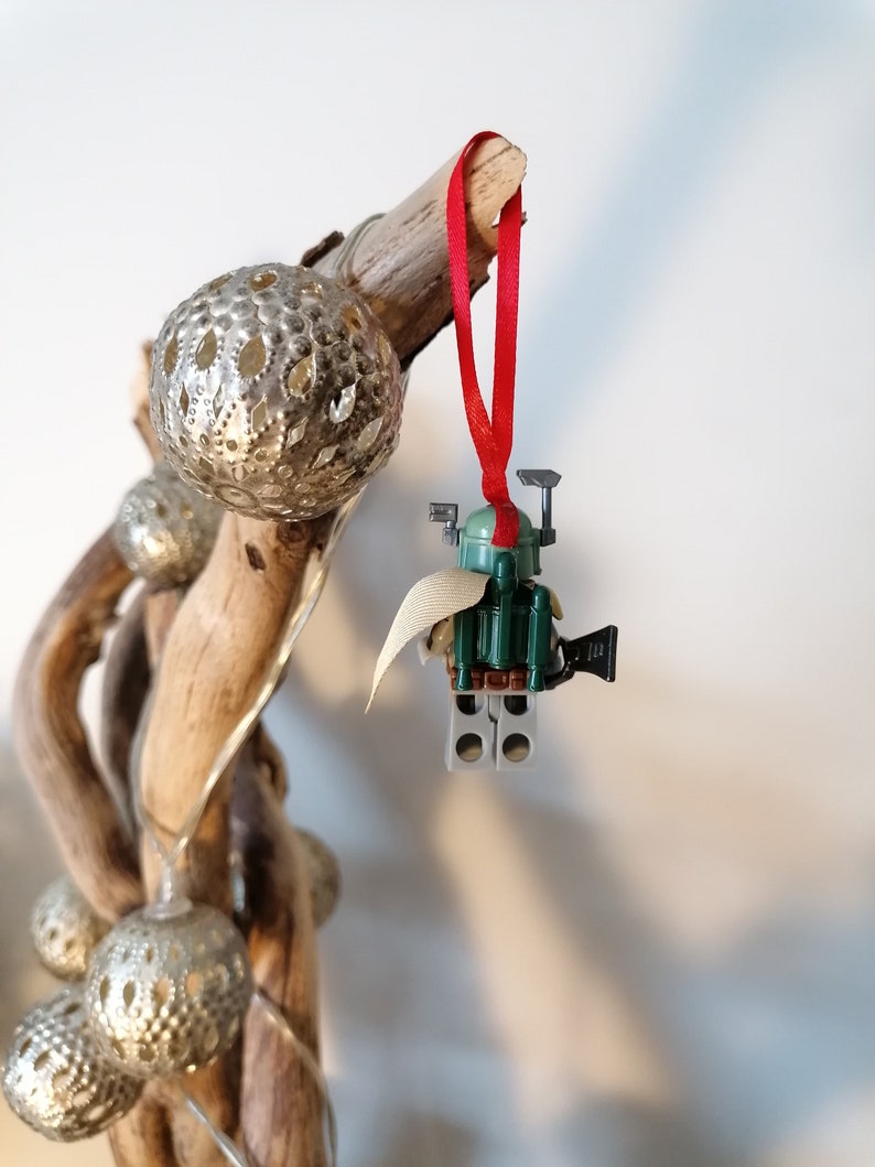 May include: A green and brown Lego Star Wars Boba Fett ornament with a red ribbon hanging from a branch with silver ornaments.