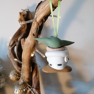 K&ouml;nnte beinhalten: Ein gr&uuml;ner und wei&szlig;er Weihnachtsbaumschmuck mit einer Figur aus dem Star Wars-Franchise. Der Schmuck h&auml;ngt an einem gr&uuml;nen Band an einem St&uuml;ck Treibholz.